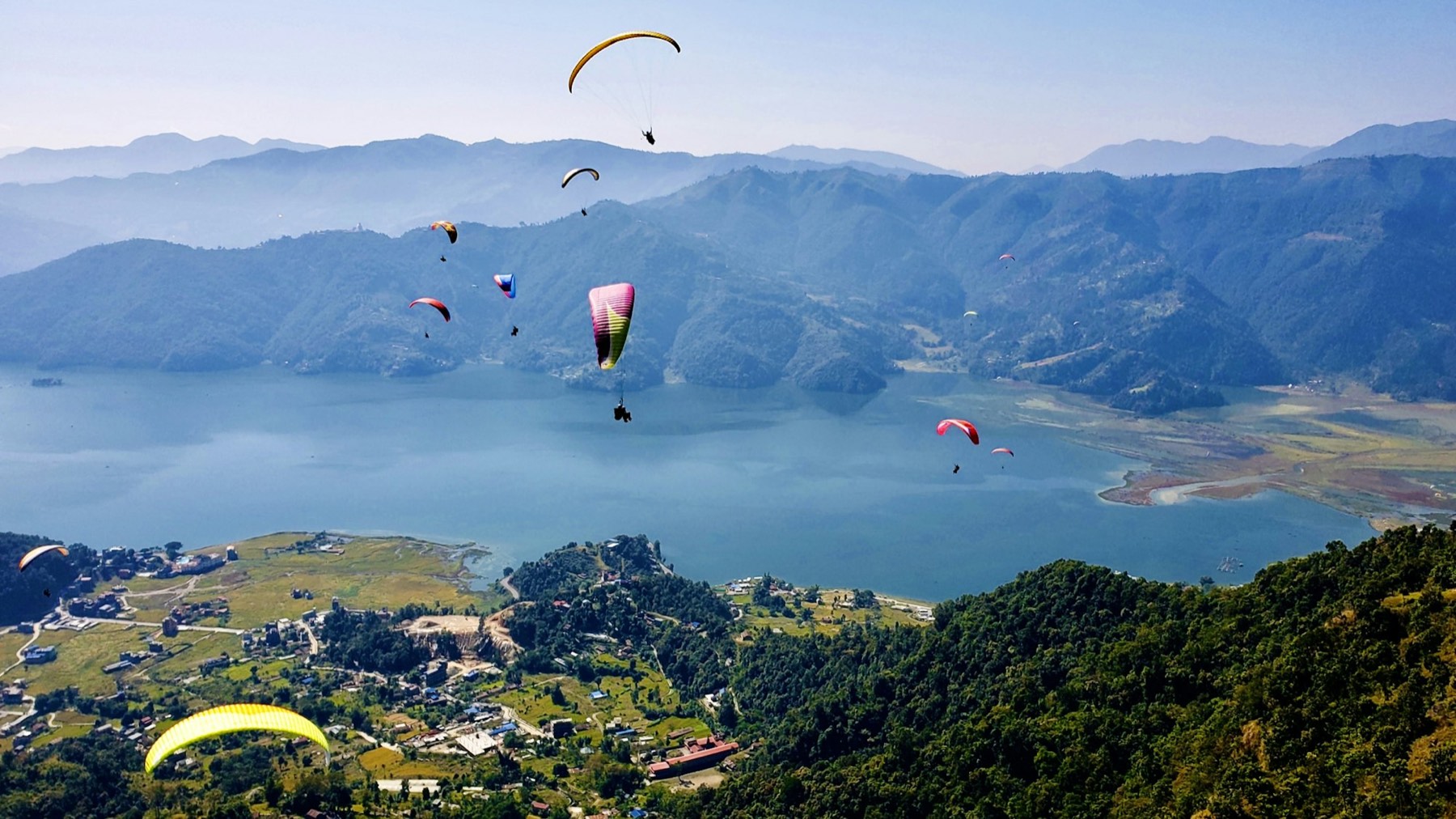 Paragliders flying above Phewa Lake, with forested ridges and a town below in Pokhara
