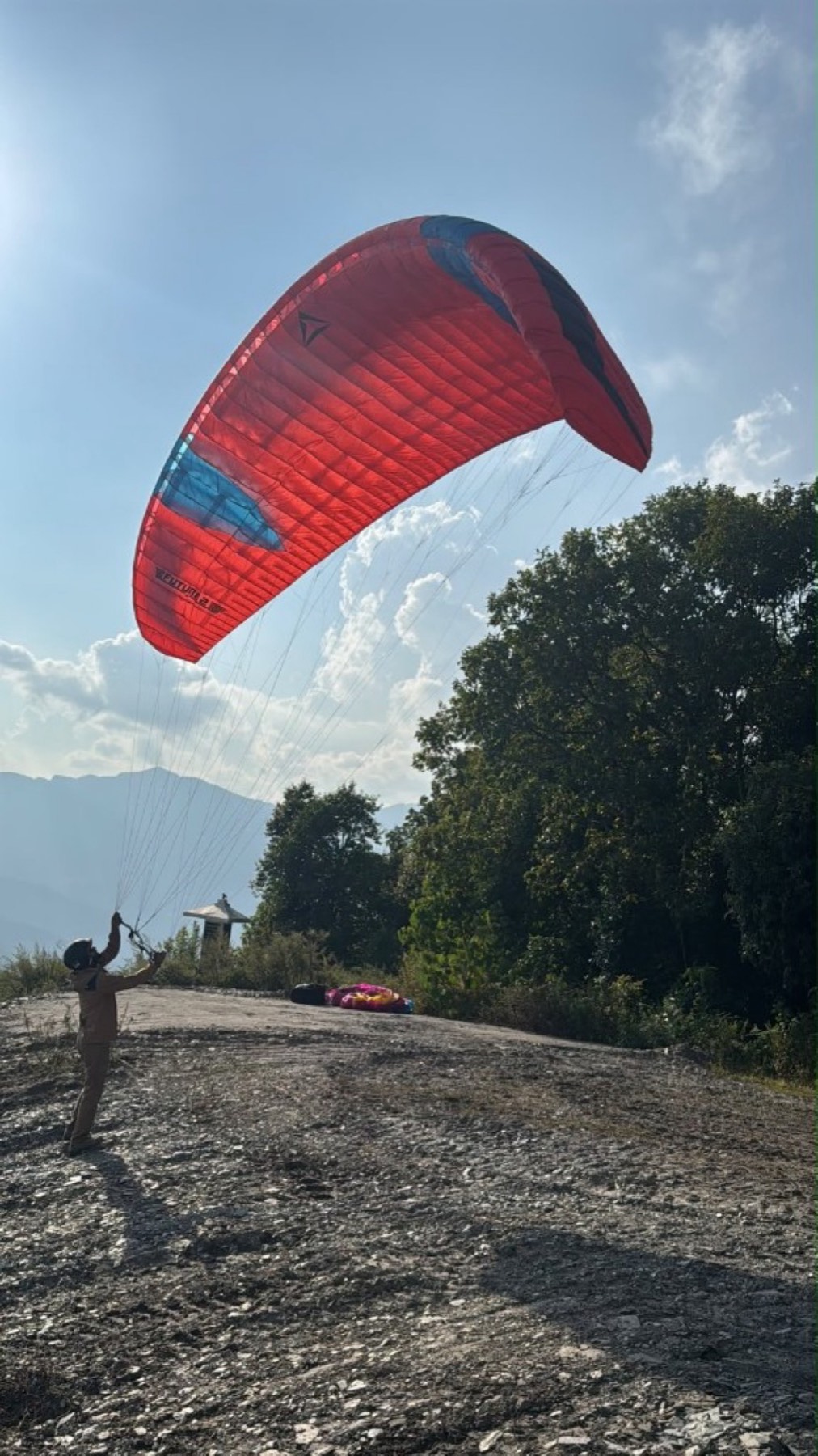 A person ground-handles a red paraglider on a rocky hillside, with trees and distant mountains